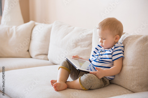 Adorable blond toddler boy laying on the sofa and playing with tablet pc at home, indoors. Child with tablet computer.