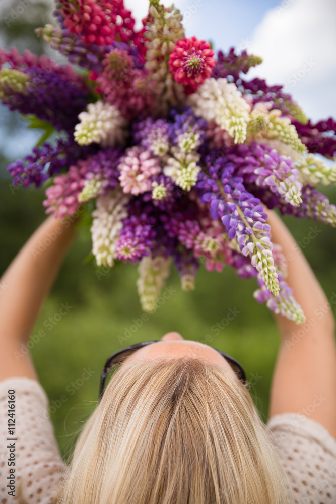 Fototapeta premium Back view on young girl holding a bunch of beautiful lupine flowers. Purple and pink lupin bouquet. Womanl with bouquet of wild lupin flowers on sunny summer field. Sky background.
