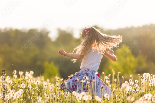 Fototapeta Naklejka Na Ścianę i Meble -  Girl running on the field of dandelions on summer sunset. Beautiful little kid girl  dancing on dandelion meadow with summer sunset. Summer fun outdoors.