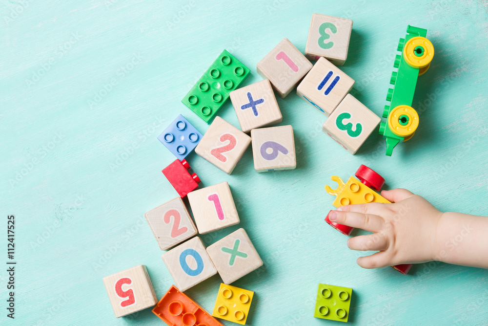 Child playing with wooden cubes with numbers and colorful toy bricks on a turquoise wooden background. Toddler learning numbers. Hand of a child taking toys. Child playing with wooden cubes with numbers and colorful toy bricks on a turquoise wooden background. Toddler learning numbers. Hand of a child taking toys.
