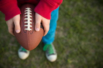 Top view on hands of little kid boy holding football on the stadium on a sunny day. Child  ready to throw a football. Sport concept. Sport activities for children outdoors.