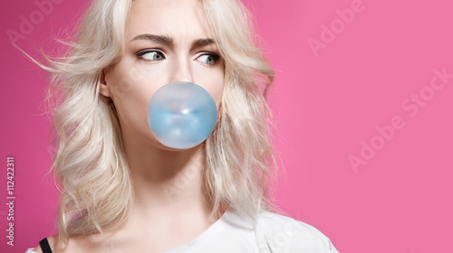 Blonde girl inflates a bubble of gum blue on a pink background