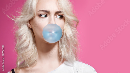 Fotografie Blonde girl inflates a bubble of gum blue on a pink background