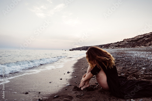 girl in black dress sitting hugging her knees on the beach at sunset