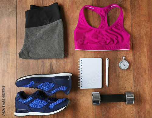 Athlete's set with female clothing, equipment and notebook on wooden background