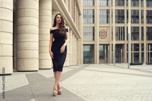Young elegant girl posing at city street. Pretty beautiful business woman in elegant black dress against city background. Full length horizontal portrait. © Dmitry Tsvetkov