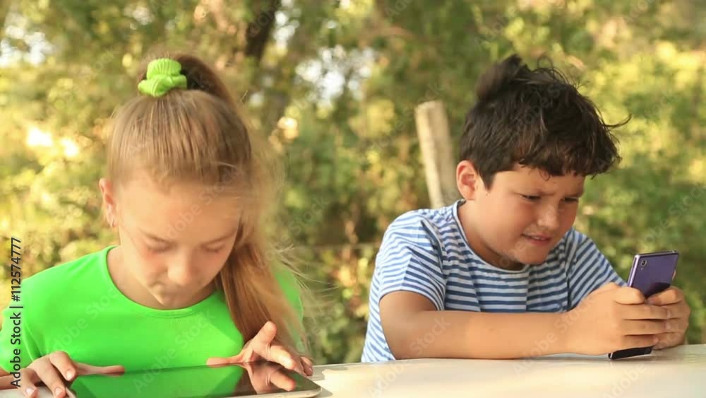 Closeup outdoor portrait of a cute young girl and boy in casual clothes sitting on the park bench with digital tablet and smart phone in her hands Closeup outdoor portrait of a cute young girl and boy in casual clothes sitting on the park bench with digital tablet and smart phone in her hands