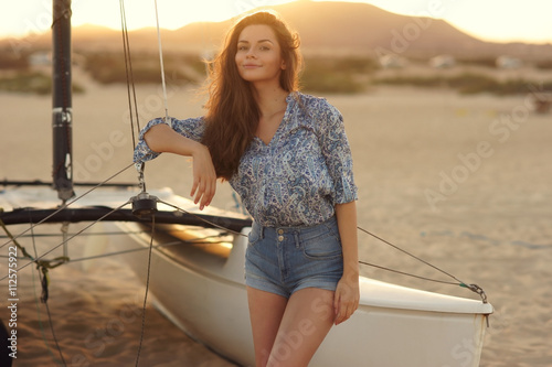 Young beautiful pretty girl sitting at sailing catamaran standing at sandy beach. Outdoor summer portrait with lovely soft evening sunset backlight