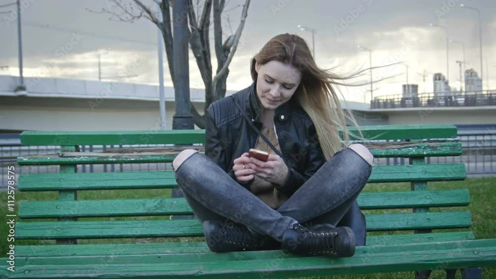 Cute happy girl with long blonde hair in leather jacket straightens hair use gadget sitting on the bench in the wind 4k Cute happy girl with long blonde hair in leather jacket straightens hair use gadget sitting on the bench in the wind 4k