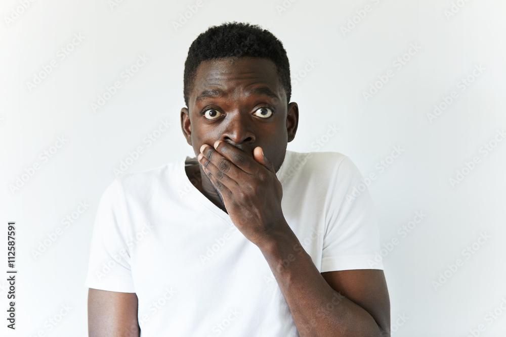 Foto de Portrait of young dark-skinned student or employee looking at the  camera with surprised guilty expression, doing something wrong, covering  his mouth. Human face expressions and emotions. Body language do Stock