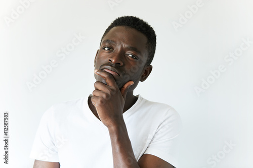 Handsome African man in white T-shirt, looking at the camera with thoughtful skeptical expression, holding hand on his chin. Black male with mistrustful look posing against white concrete wall