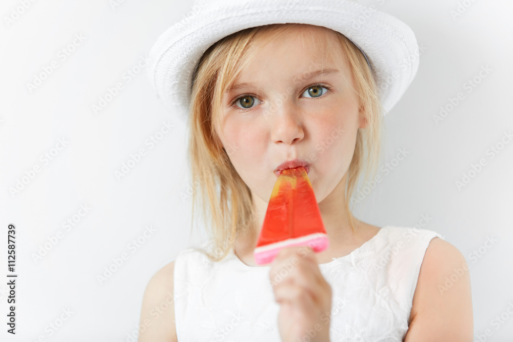 Little Caucasian girl eating her popsicle in white dress and white summer hat indoors. Sweet tooth is glad to enjoy her yum yum and watching straight forward in relaxed manner.