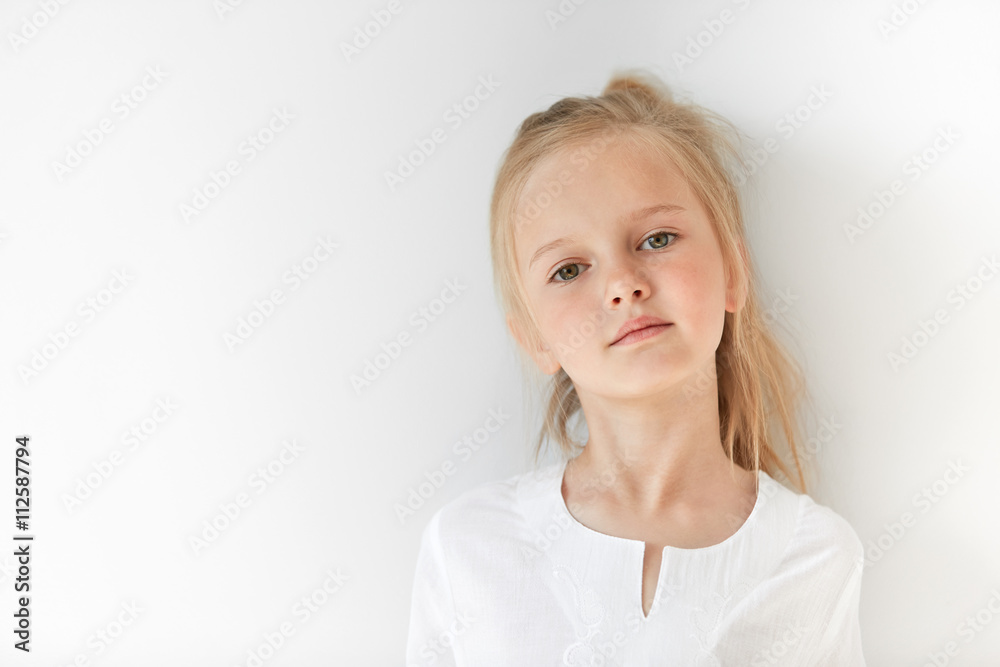 Small girl in white light room looking pretentiously at camera with her  chin raised and head tilted. Pride and honor mixed with childish  self-assurance and confidence show her superior attitude. Stock Photo