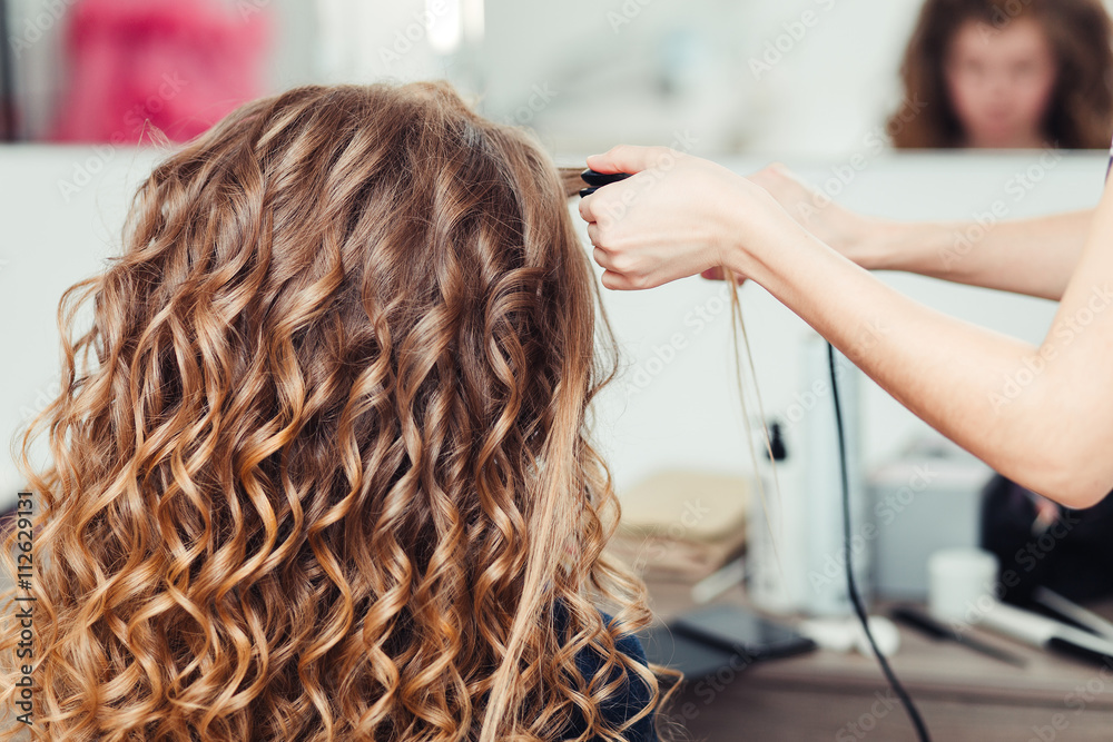 Fototapeta premium Hairdresser curling woman hair with electric iron curler tong. Hairstylist making girl hairstyle.