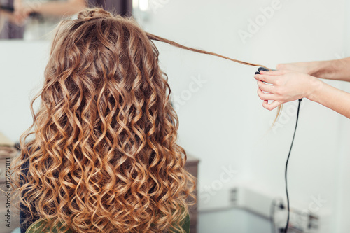 фотографія Hairdresser curling woman hair with electric iron curler tong