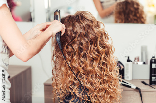 Hairdresser curling woman hair with electric iron curler tong. Hairstylist making girl hairstyle.