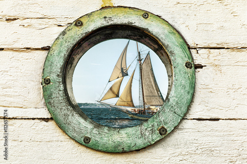 Old porthole window looks out at an old sailing ship. Vintage color