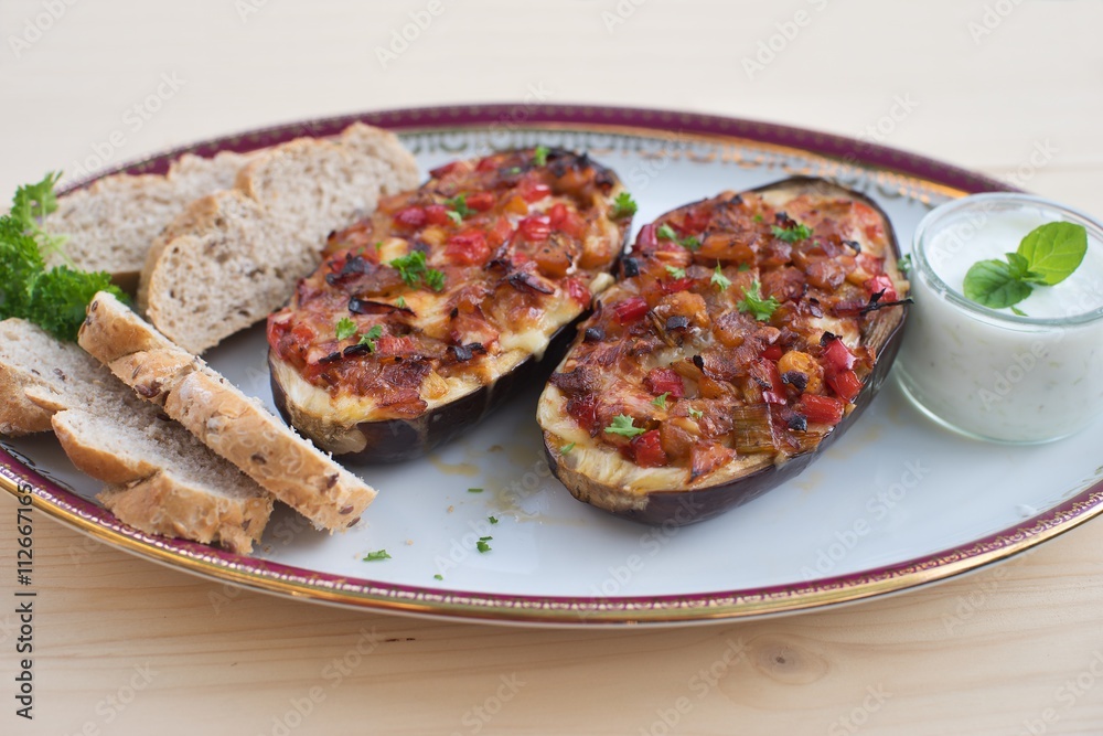 Stuffed Eggplant halves with tomato onion sweet pepper and cheese baked and served with small pieces of whole grain bread roll and greek style sauce made from yogurt, cucumber, garlic and mint leaves