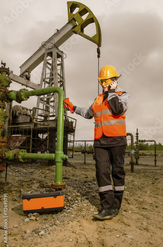 Woman engineer in the oil field talking on the radio wearing orange helmet and work clothes. Pump jack background. Oil and gas concept. Toned.