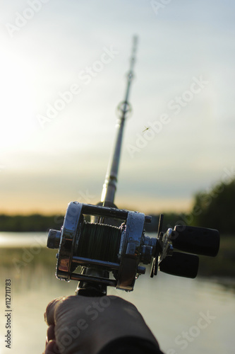 Male hand holding a round fishing reel attached to a fishing rod with blurred sea nature on the background at sunset time.  photo