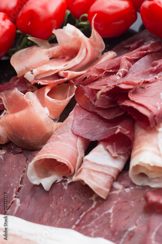 Jambon mix. Ham. Traditional Italian and Spanish salting, smoking, dry-cured dish - jamon Serrano and prosciutto crudo sliced with herbs and tomatos on dark stone background. Copy space. Closeup. 
