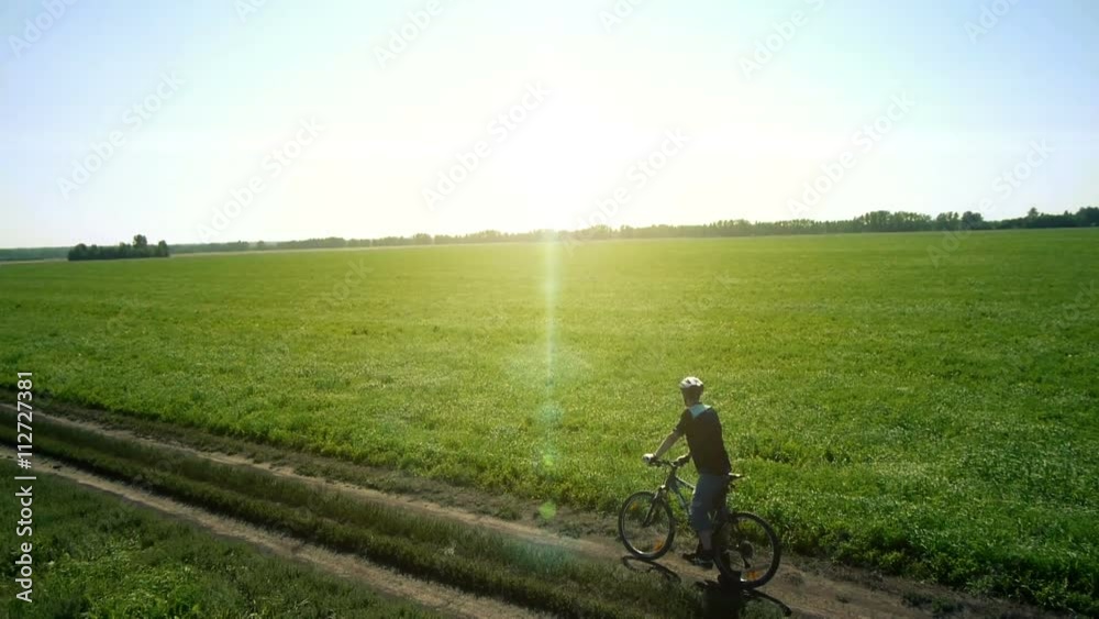AERIAL: Young man cycling on bicycle at rural road through green and yellow summer meadow field. During sunny day. AERIAL: Young man cycling on bicycle at rural road through green and yellow summer meadow field. During sunny day.