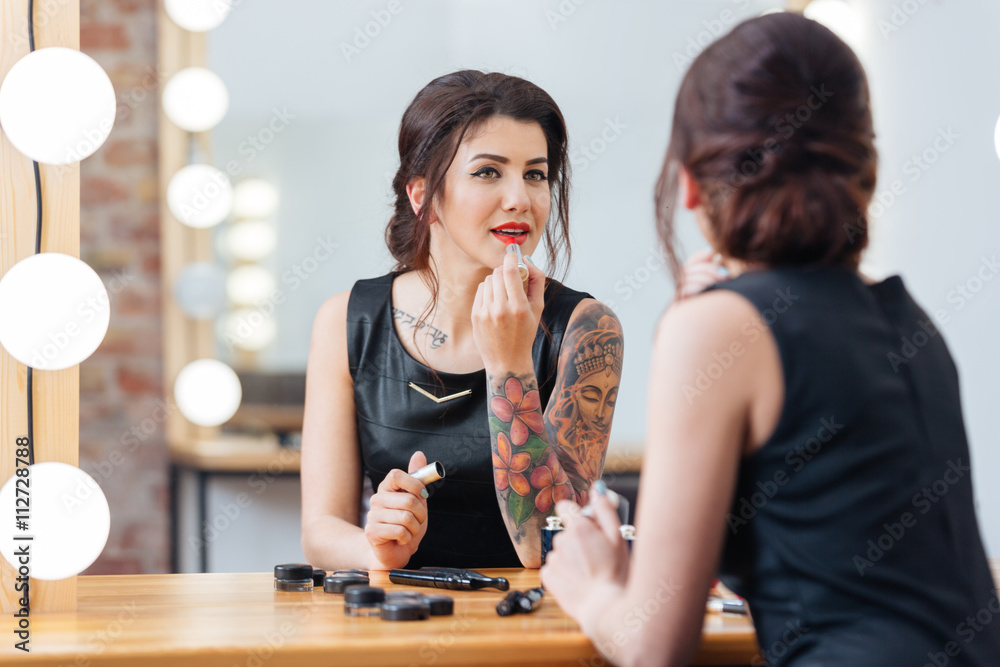 Fototapeta premium Beautiful tattooed woman applying lipstick in dressing room