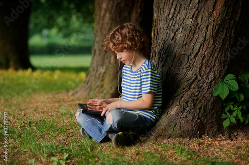 The boy with the tablet in hands sits under a big tree.