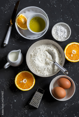 Ingredients for making orange cake with olive oil - flour, eggs, olive oil, powdered sugar on a dark stone background