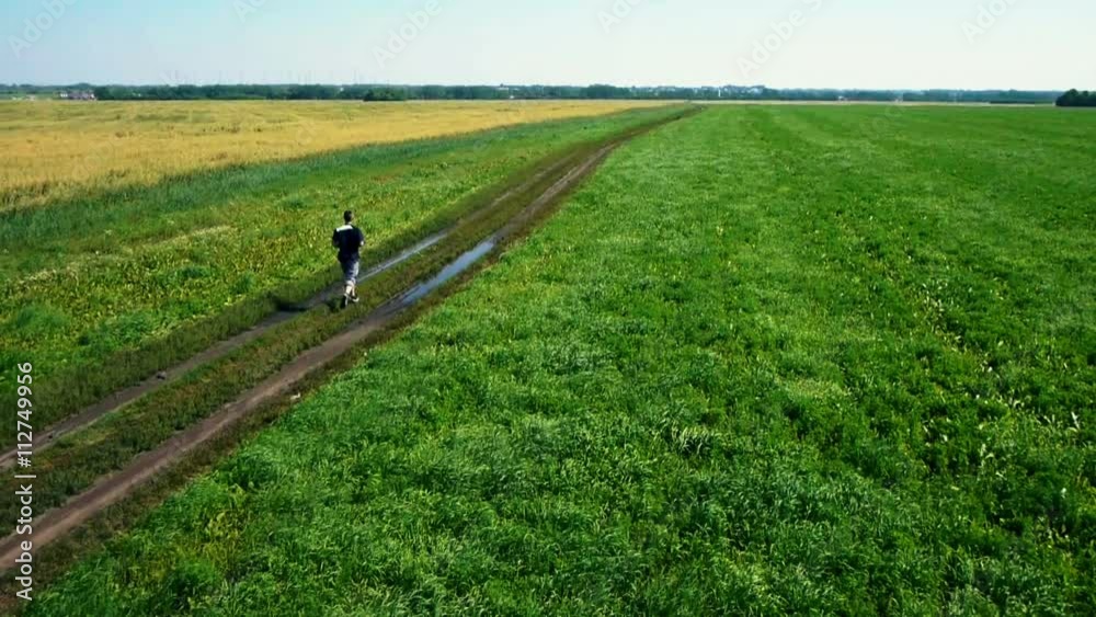 AERIAL: Running athlete man. Male runner jogging during outdoors training. Athletic fit young sport fitness model on rural road outside in green field. AERIAL: Running athlete man. Male runner jogging during outdoors training. Athletic fit young sport fitness model on rural road outside in green field.