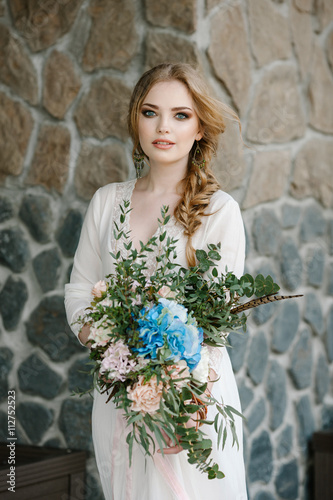 невеста в белом платье держит в руках большой букет в стиле бохо на фоне красивой стены из дикого камня серого и бежевого цвета