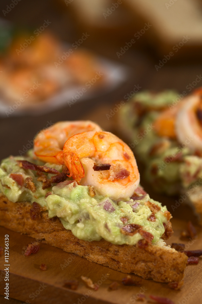 Vollkorntoastbrot belegt mit Guacamole (Avokado-Creme), gebratenen Garnelen und Speckstückchen (Selektiver Fokus, Fokus auf die Vorderseite der Garnele auf dem Brot)