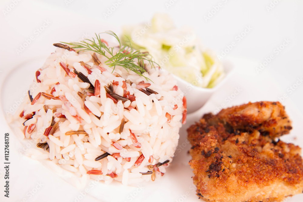 Pork chop (covered in breadcrumbs), three color rice and cucumber salad decorated with dill on a plate on white background