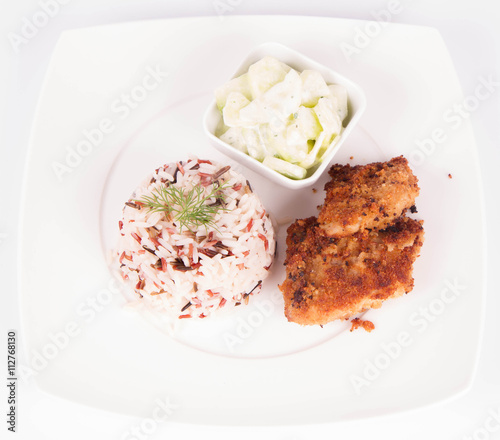 Pork chop (covered in breadcrumbs), three color rice and cucumber salad decorated with dill on a plate on white background