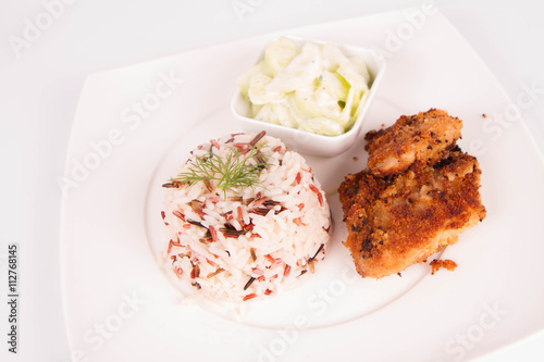 Pork chop (covered in breadcrumbs), three color rice and cucumber salad decorated with dill on a plate on white background