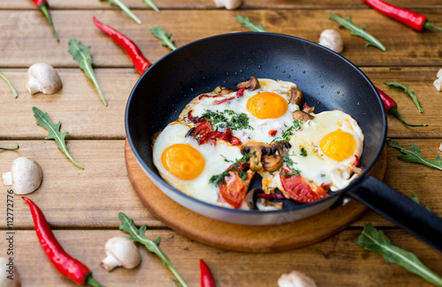 Breakfast set. Pan of fried eggs with mushrooms, fresh tomato, on rustic serving board over wooden background