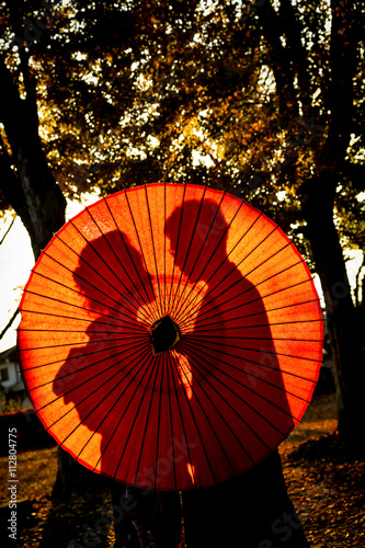 Traditional Japanese ceremony wedding lovely day  silhouettes of married couple holding red paper umbrella in hands  kissing under golden sunset in shrine temple garden  colorful maple ginkgo leaves