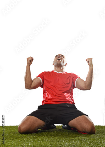 happy and excited football player in red jersey celebrating scoring goal kneeling on grass pitch