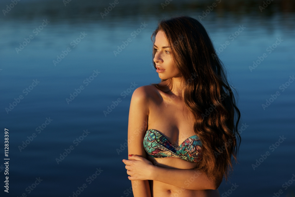 Fototapeta premium Young beautiful pretty woman standing and posing in blue bikini in water in sunset soft lightning. Fashion style outdoor summer portrait