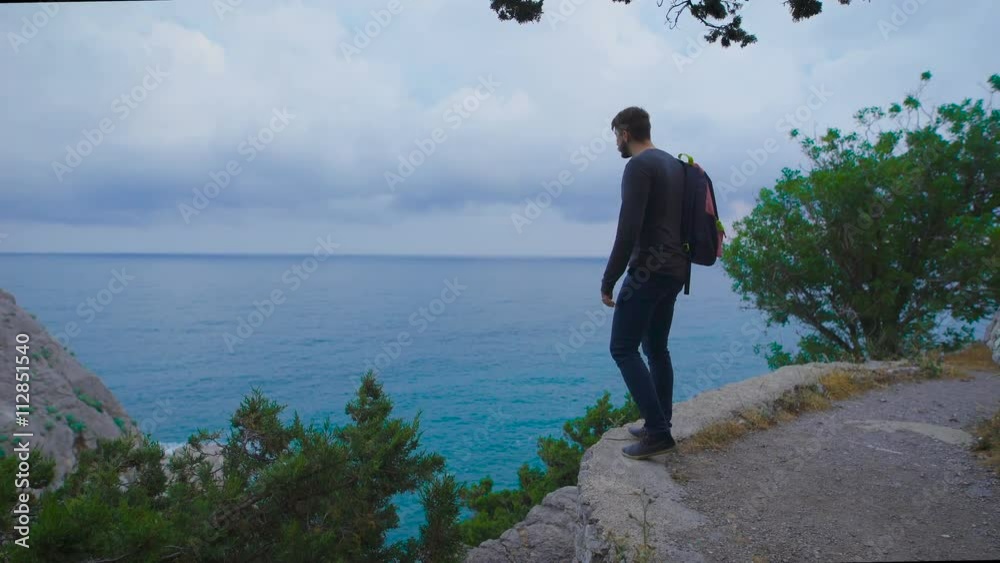 Young hipster with backpack suitable to the edge and looks at the stunning views. Young hipster with backpack suitable to the edge and looks at the stunning views.