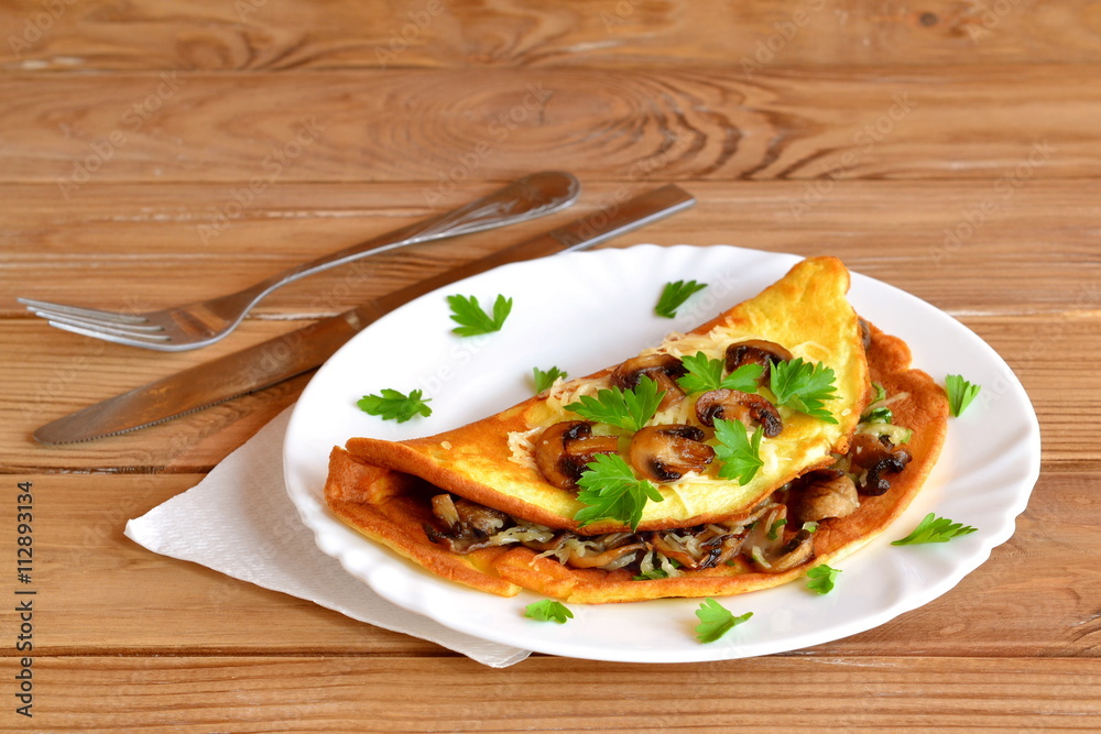 Fried omelette with mushrooms, cheese and green parsley. Stuffed omelette on a plate and on a wooden table. Fork, knife, napkin. Breakfast eggs recipe. Breakfast idea
