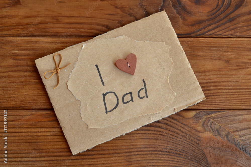 Homemade greeting card on wooden table. Card with text I love dad. Card is made by hand from cardboard box, wrapping paper, wooden button, cord. Children gift for father. Easy home present idea. Homemade greeting card on wooden table. Card with text I love dad. Card is made by hand from cardboard box, wrapping paper, wooden button, cord. Children gift for father. Easy home present idea.