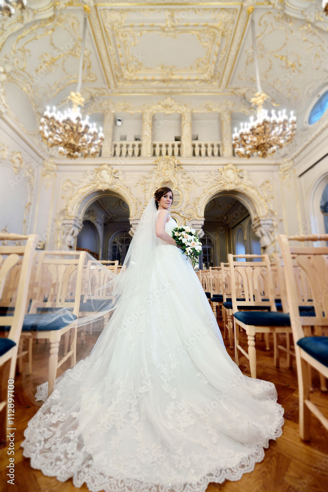 Beauty bride in bridal gown with bouquet and lace veil indoors. Beautiful model girl in a white wedding dress. Female portrait of cute lady. Woman with hairstyle