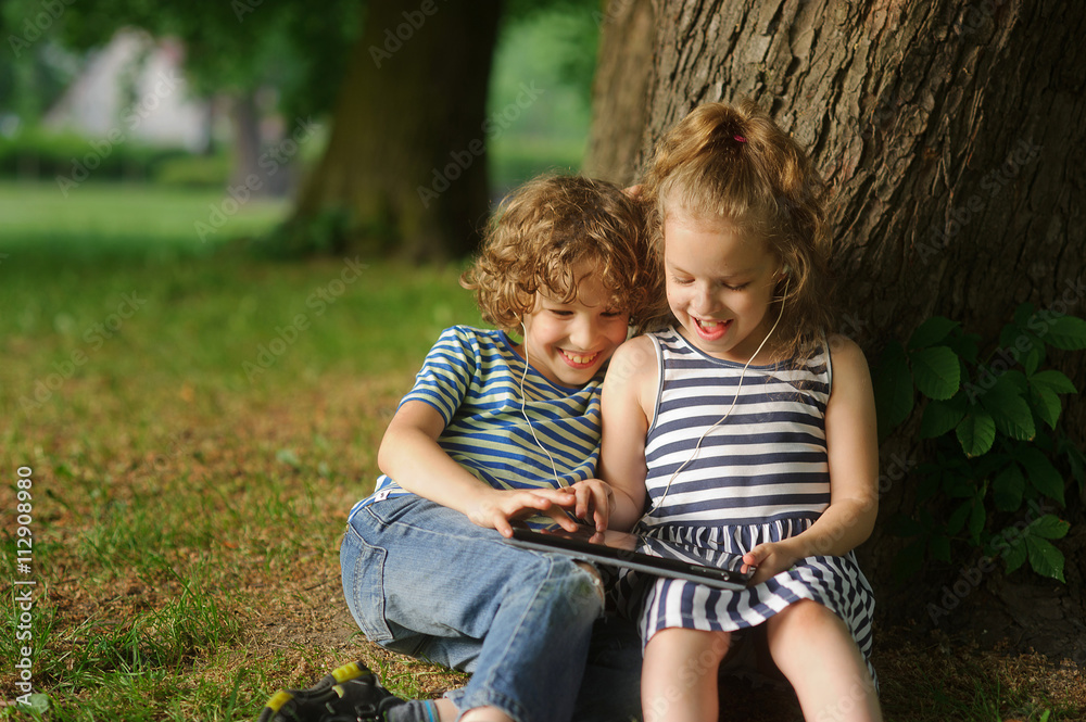 Two children sit in park under a big tree and play on the tablet. Two children sit in park under a big tree and play on the tablet.