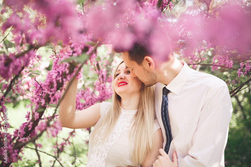 Young beautiful couple kissing and hugging near trees with blossom in summer park Young beautiful couple kissing and hugging near trees with blossom in summer park