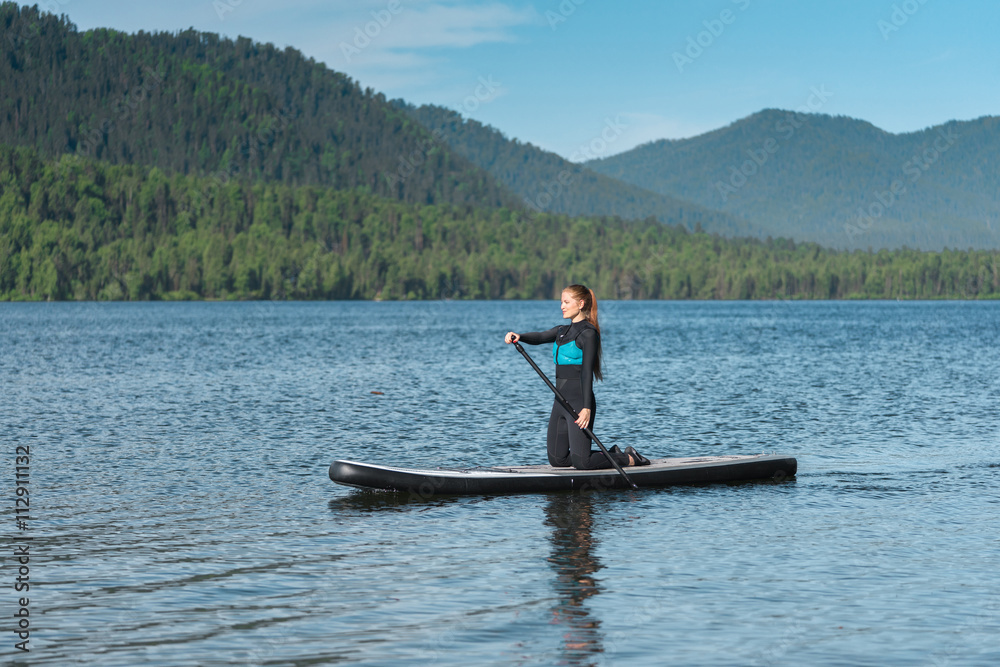 Smiling woman paddle boarding on knees at the mountain lake 