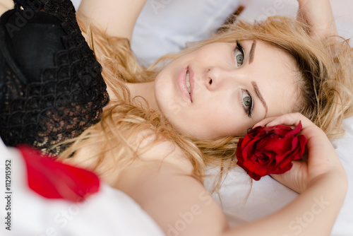 closeup portrait of beautiful young lady having fun holding rose flower. romantic pinup silk skin female relaxing in bed on light copy space background and looking at camera