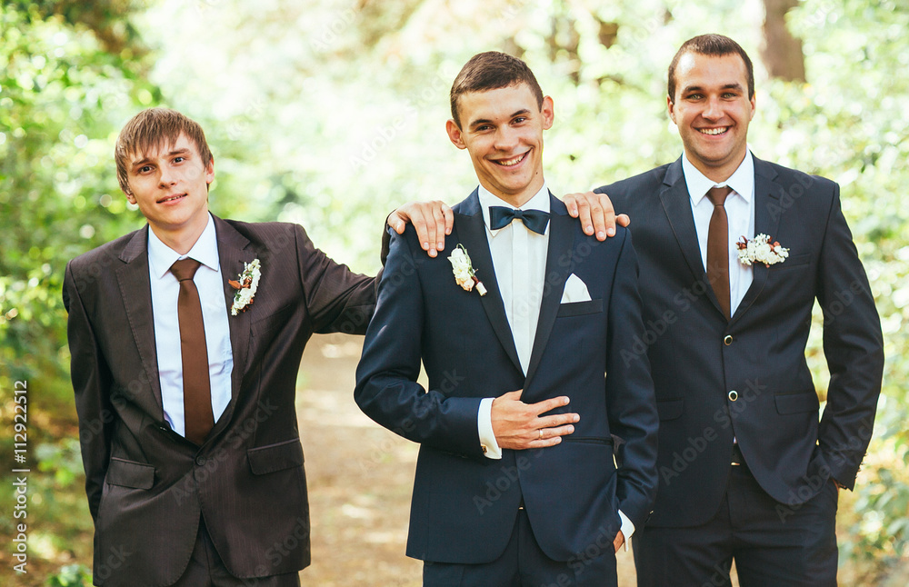 Confident smiling handsome groom in black suit with two groomsman in pine wooden park