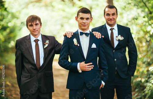 Confident smiling handsome groom in black suit with two groomsman in pine wooden park