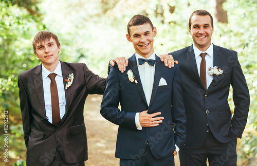 Obraz na płótnie Confident smiling handsome groom in black suit with two groomsman in pine wooden park w salonie